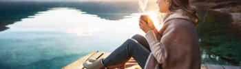 Mladá žena sedí u jezera s hrnkem kávy a relaxuje, aby uklidnila mysl a snížila stres