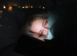 Tipy pro lepší spánek: žena unavená spí v posteli, osvícená světlem mobilního telefonu.