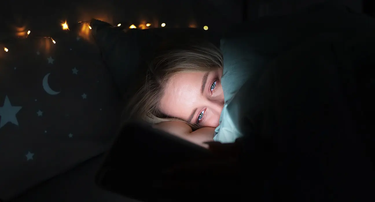 Tipy pro lepší spánek: žena unavená spí v posteli, osvícená světlem mobilního telefonu.