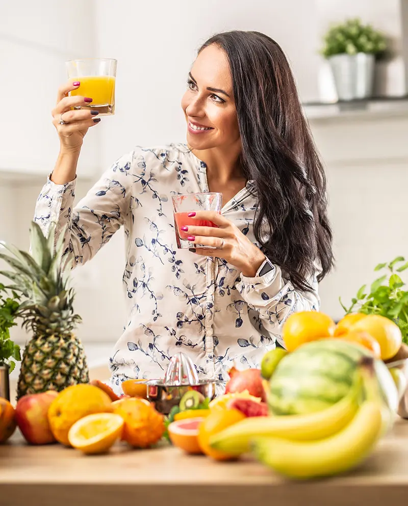 Mladá žena v kuchyni drží sklenici fresh juice, obklopená ovocem bohatým na vitamín C pro posílení imunity.